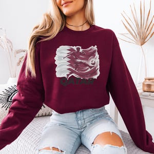 A woman wearing a burgundy sweatshirt with a graphic of a bird and the word "QATAR" printed on it, sitting on a bed with a white pillow.