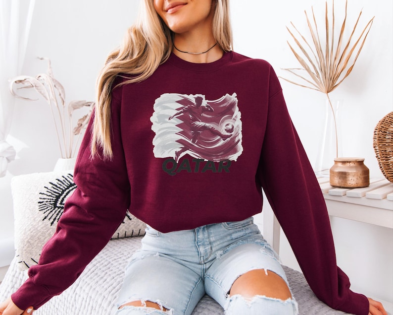 A woman wearing a burgundy sweatshirt with a graphic of a bird and the word "QATAR" printed on it, sitting on a bed with a white pillow.