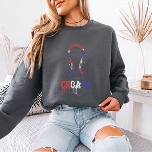 A woman wearing a grey sweatshirt with a graphic of the European Cup logo, sitting on a couch in a room with a plant and a vase.
