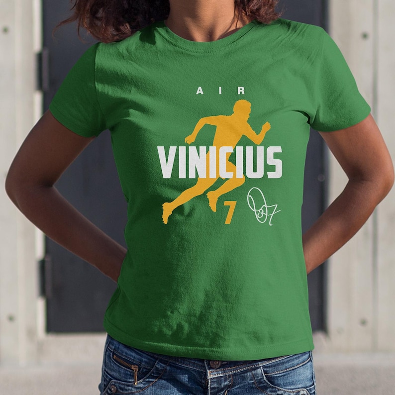 A woman wearing a green t-shirt with a graphic design featuring a yellow figure and the text "AIR VINICius 7" printed on it.