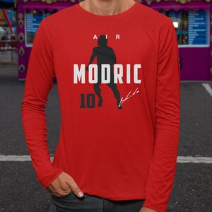 A man wearing a red long-sleeved shirt with a graphic design featuring a silhouette of a person and the text "MODRIC 10" printed on it.