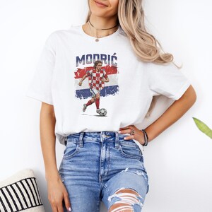 A woman wearing a white t-shirt with a graphic of a soccer player and blue jeans stands in front of a plant.