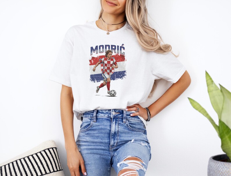 A woman wearing a white t-shirt with a graphic of a soccer player and blue jeans stands in front of a plant.
