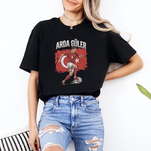 May include: Black t-shirt featuring the text "ARDA GÜLER" above an illustration of a soccer player in a red and white uniform kicking a soccer ball. The illustration is set against a distressed Turkish flag.