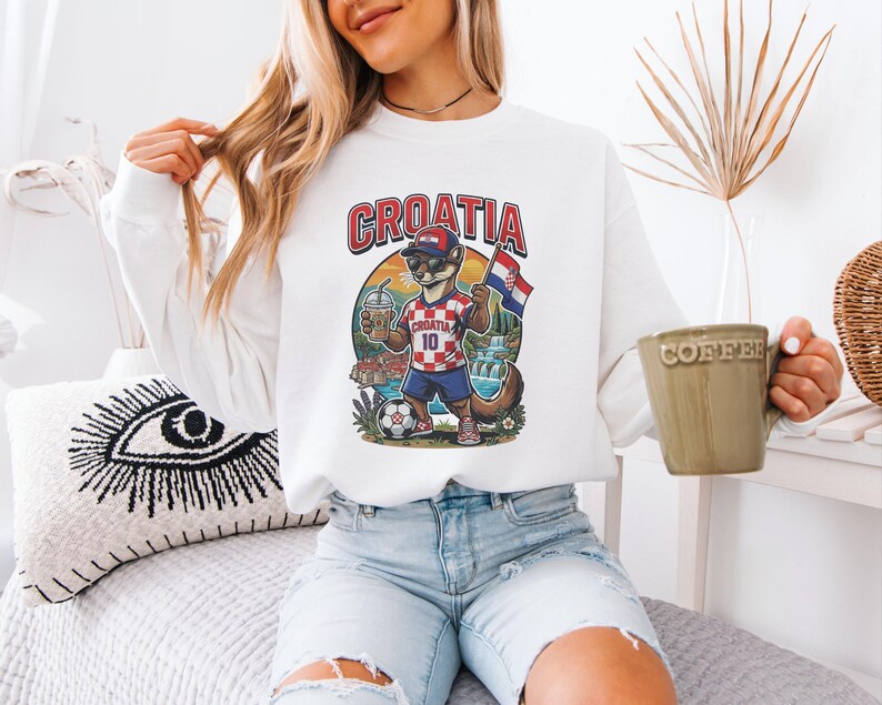 A woman wearing a white sweatshirt with a graphic of a cat holding a soccer ball and a flag, sitting on a bed and holding a coffee mug.