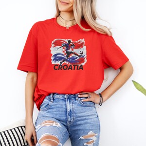 A woman wearing a red t-shirt with a graphic of a soccer player and the word "Croatia" printed on it, standing in front of a white wall.