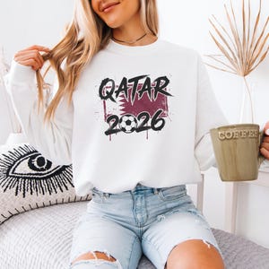 A woman wearing a white sweatshirt with the text "QATAR 2026" and holding a coffee mug is sitting on a bed.