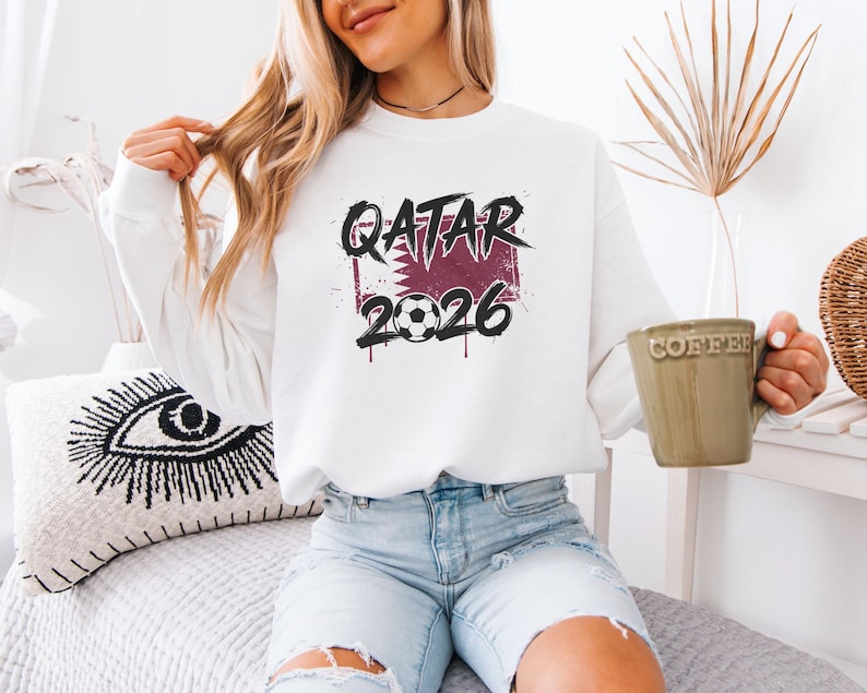 A woman wearing a white sweatshirt with the text "QATAR 2026" and holding a coffee mug is sitting on a bed.