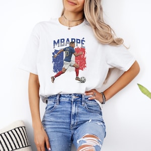 A woman wearing a white t-shirt with a graphic of a soccer player and blue jeans stands in front of a plant.