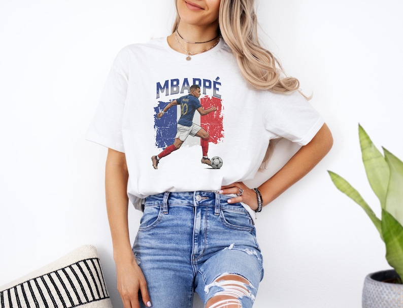 A woman wearing a white t-shirt with a graphic of a soccer player and blue jeans stands in front of a plant.