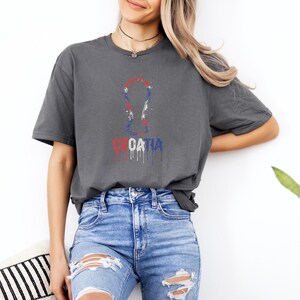 A woman wearing a gray t-shirt with a graphic of the European Cup and the word "Croatia" printed on it, standing against a white wall.