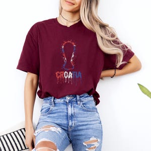 A woman wearing a burgundy t-shirt with a graphic of a trophy and the word "Croatia" printed on it, paired with distressed blue jeans.