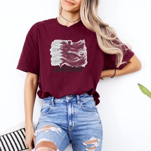A woman wearing a burgundy t-shirt with a graphic of a pig and blue jeans stands against a white wall, with a potted plant visible in the background.