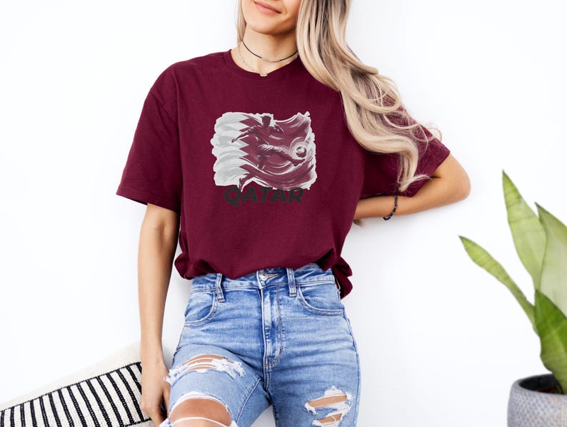 A woman wearing a burgundy t-shirt with a graphic of a pig and blue jeans stands against a white wall, with a potted plant visible in the background.