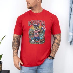 A man wearing a red t-shirt with a graphic of a dog holding a soccer ball and a flag, standing in front of a white wall.