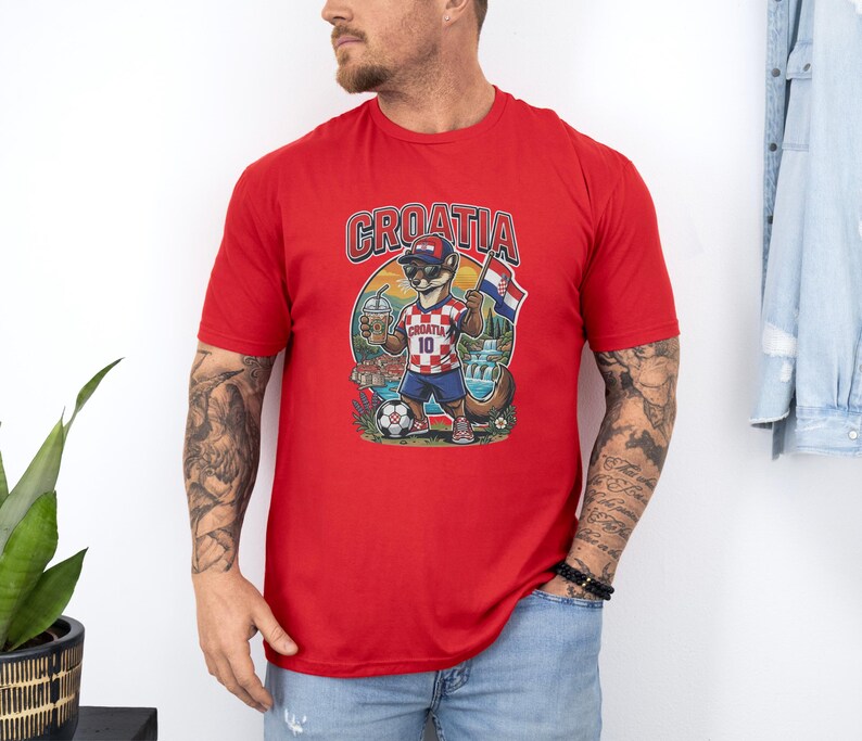 A man wearing a red t-shirt with a graphic of a dog holding a soccer ball and a flag, standing in front of a white wall.
