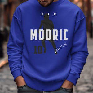 A man wearing a blue sweatshirt with a graphic design featuring a silhouette of a person and the text "MODRIC 10" printed on it.