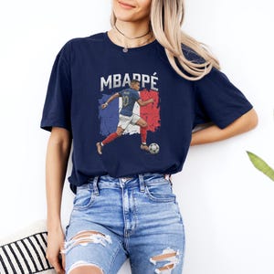 A woman wearing a blue t-shirt with a graphic of a soccer player and the text "MBARPE" is standing against a white wall.