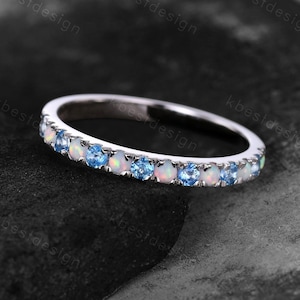 Può includere: Un anello d'argento con una fascia di pietre blu e opali bianchi alternati. L'anello è posto su uno sfondo scuro e strutturato, che mette in risalto la brillantezza delle pietre. L'anello è un gioiello.