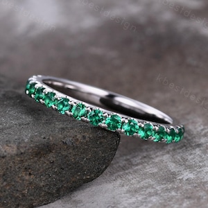 Peut inclure: Une bague en argent ornée d'une rangée continue de petites pierres précieuses vertes vives. La bague est sertie de pierres vertes émeraude, créant un effet scintillant. La bague est présentée sur un fond sombre et texturé.