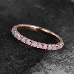 May include: Rose gold ring featuring a band of small, round, white opal stones. The ring is set against a dark, textured background, emphasising the delicate design and the subtle iridescence of the opals.