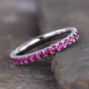 May include: A silver ring with a band of vibrant pink gemstones. The ring is set against a textured gray surface, highlighting the sparkle of the gemstones. The ring is a piece of jewelry.