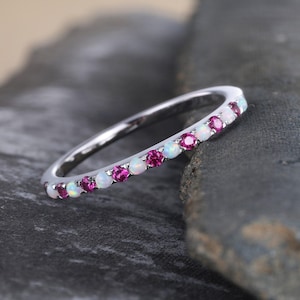 May include: A silver ring with alternating pink and white gemstones. The ring is set on a dark gray stone surface. The gemstones are evenly spaced around the band, creating a continuous pattern.