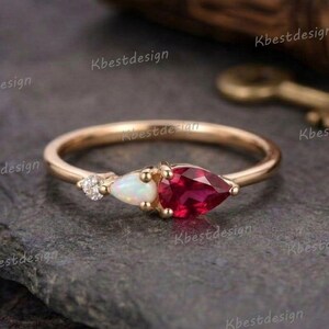 Könnte beinhalten: Ein goldener Ring mit einem kleinen Diamanten, einem ovalen Opal und einem birnenförmigen Rubin. Der Ring liegt auf einer dunkelgrauen Oberfläche. Der Ring ist ein Schmuckstück.
