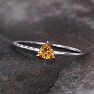 Puede incluir: Un delicado anillo de plata con una gema triangular de color amarillo dorado. La gema es el punto focal, engastada en una fina banda pulida. El diseño del anillo es minimalista y elegante.