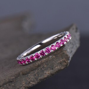 Puede incluir: Un anillo plateado con una banda de peque&ntilde;as y vibrantes piedras rosas. El anillo se presenta sobre un fondo oscuro y texturizado, resaltando el brillo de las piedras. Una pieza de joyer&iacute;a cl&aacute;sica y elegante.