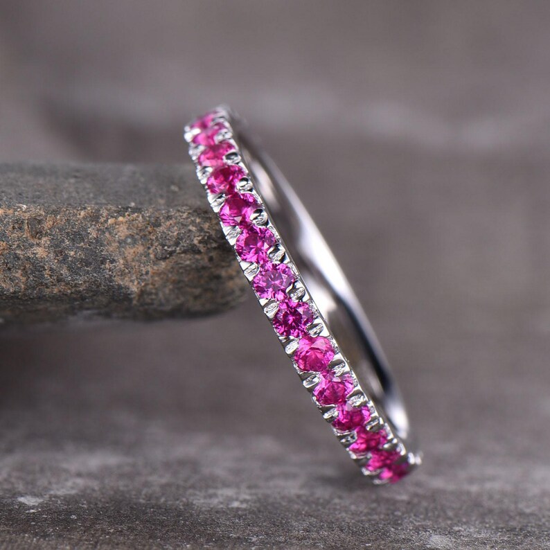 Puede incluir: Un anillo de plata con una banda de gemas rosas vibrantes. El anillo se muestra sobre un fondo gris oscuro y texturizado, resaltando el brillo de las gemas. El dise&ntilde;o del anillo es simple y elegante.