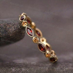 May include: A gold ring with alternating oval and round gemstones. The ring features deep red and golden-yellow stones set in a repeating pattern. The ring is displayed on a dark, textured surface.