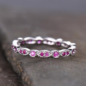 Può includere: Un anello d'argento con una fascia continua di piccole gemme rosa rotonde. L'incastonatura dell'anello presenta un delicato design smerlato e le gemme sono uniformemente distanziate attorno alla fascia. L'anello è esposto su una superficie scura e strutturata.