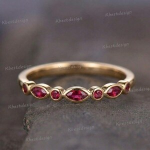 May include: A gold-colored ring with alternating round and marquise-cut red gemstones. The ring is set against a blurred background. The text "Kbestdesign" is repeated around the ring.