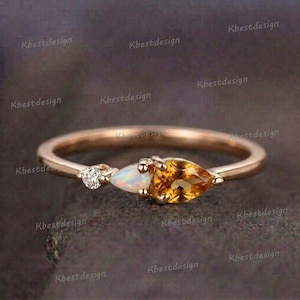 May include: A gold ring featuring three gemstones: a small diamond, an opal, and a pear-shaped citrine. The ring has a thin band and is set against a dark, textured background. The word "Kbestdesign" is repeated around the ring.