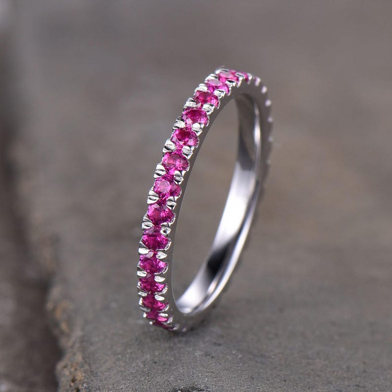 Puede incluir: Un anillo plateado con una hilera continua de gemas de color rosa brillante. El anillo se muestra sobre un fondo gris neutro, acentuando el color de las gemas. Una joya atemporal y elegante.