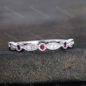 Puede incluir: Un delicado anillo de plata con piedras preciosas rojas y claras alternas. El anillo presenta piedras rojas redondas y piedras claras ovaladas. El anillo tiene una banda delgada y un patrón repetitivo de piedras preciosas. El anillo está sobre una superficie oscura y texturizada.