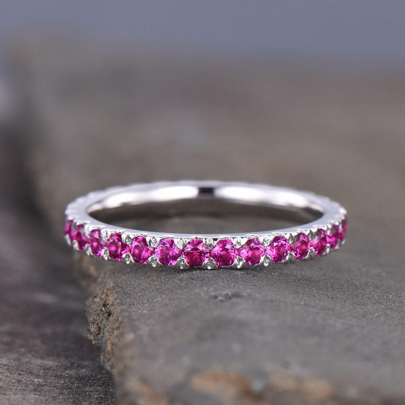Puede incluir: Un anillo de plata con una fila continua de peque&ntilde;as piedras preciosas rosas brillantes. El anillo se muestra sobre una superficie gris texturizada, resaltando el brillo de las piedras. Es una pieza de joyer&iacute;a.