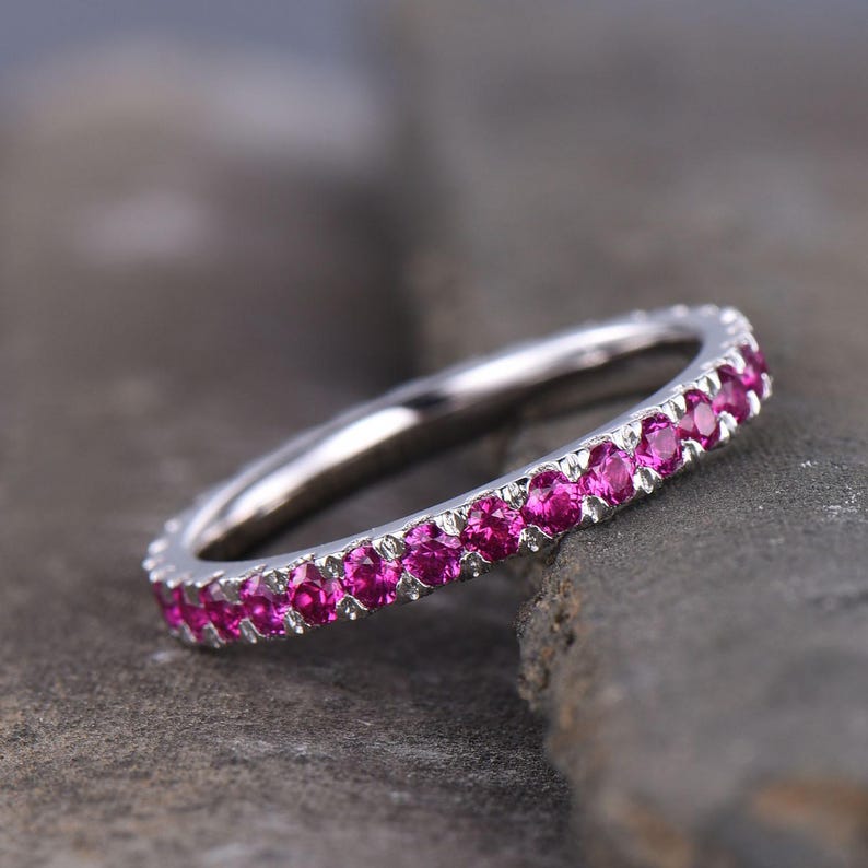 Puede incluir: Un anillo plateado con una fila continua de gemas de color rosa brillante. El anillo se muestra sobre una superficie gris texturizada, acentuando el color de las gemas. Esta es una pieza de joyer&iacute;a.