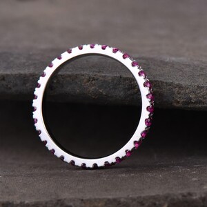 Puede incluir: Un anillo de plata con una fila continua de peque&ntilde;as piedras preciosas redondas de color rosa brillante. El anillo se encuentra sobre una superficie gris oscura y texturizada, resaltando el metal pulido y las vibrantes piedras. Es una joya.