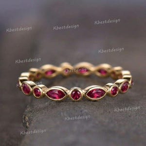 May include: A gold-colored ring featuring alternating oval and round red gemstones. The ring has a continuous design of gemstones set in a band. The ring is displayed on a dark, textured surface.