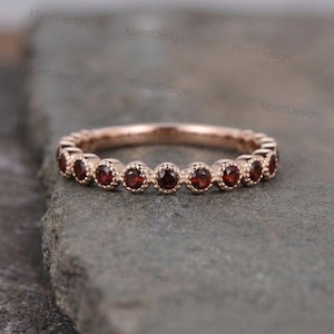 Op de afbeelding: Roségouden ring met een reeks ronde, dieprode edelstenen. De edelstenen zijn gezet in individuele bezels, waardoor een doorlopende band ontstaat. De ring wordt getoond op een getextureerd grijs oppervlak.