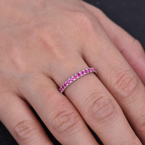 Puede incluir: Un anillo plateado con una banda de peque&ntilde;as y vibrantes piedras preciosas rosas. El anillo se lleva en un dedo, mostrando la fila continua de piedras brillantes. Las piedras preciosas est&aacute;n muy juntas, creando un efecto brillante.