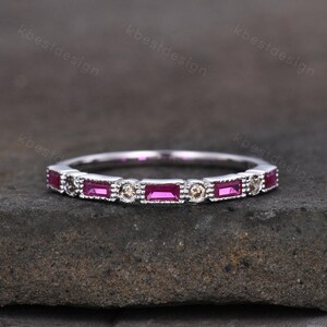 May include: A silver band ring featuring alternating rectangular and round gemstones. The rectangular stones are a vibrant magenta color, while the round stones are a light champagne color. The ring is set on a dark gray surface.