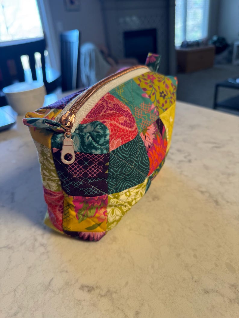 Peut inclure: Une trousse de maquillage patchwork color&eacute;e avec une fermeture &eacute;clair dor&eacute;e. Le sac pr&eacute;sente une vari&eacute;t&eacute; de motifs de tissus dans les tons de rose, vert, jaune et violet. Le sac est rectangulaire et poss&egrave;de une petite boucle en tissu.
