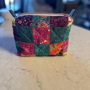 Op de afbeelding: Een vierkante, gewatteerde patchwork-etui met ritssluiting. De etui is gemaakt van een verscheidenheid aan stoffen met bloemen- en geometrische patronen in tinten van teal, paars, roze en oranje. De rits is wit met een goudkleurige ritstrekker en heeft stoffen lussen aan elke kant.