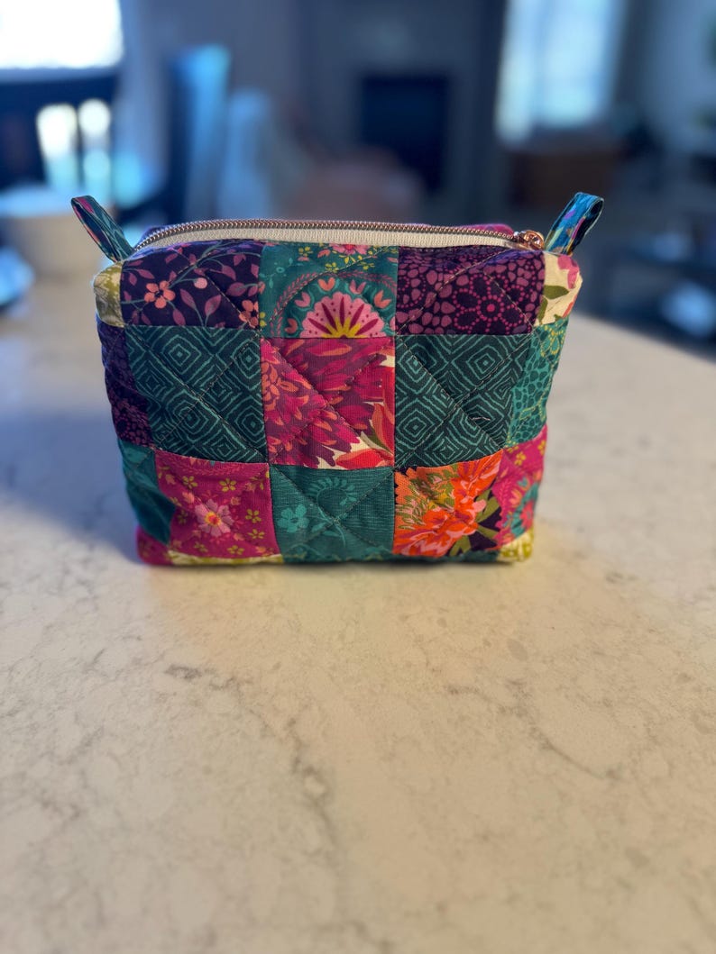Op de afbeelding: Een vierkante, gewatteerde patchwork-etui met ritssluiting. De etui is gemaakt van een verscheidenheid aan stoffen met bloemen- en geometrische patronen in tinten van teal, paars, roze en oranje. De rits is wit met een goudkleurige ritstrekker en heeft stoffen lussen aan elke kant.