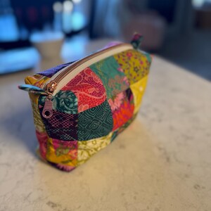 Peut inclure: Une trousse de maquillage patchwork color&eacute;e avec une fermeture &eacute;clair en or rose. Le sac pr&eacute;sente une vari&eacute;t&eacute; de motifs de tissus dans les tons de rose, vert, jaune et violet. Le sac est pos&eacute; sur une surface blanche.