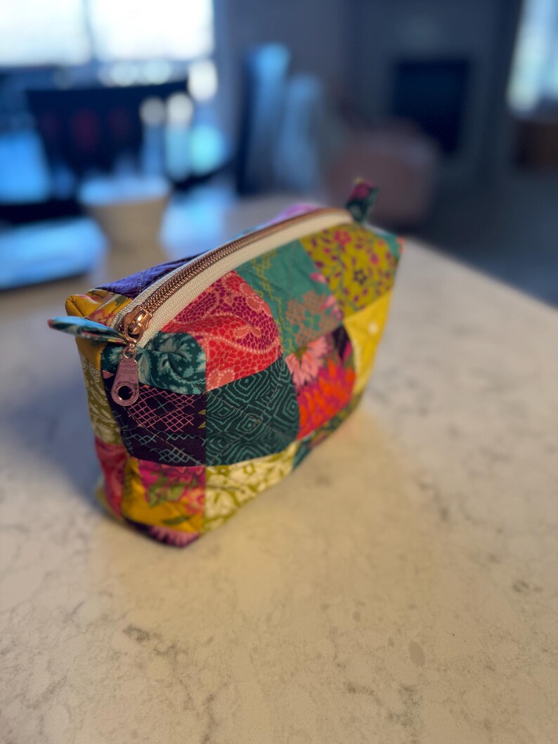 Peut inclure: Une trousse de maquillage patchwork color&eacute;e avec une fermeture &eacute;clair en or rose. Le sac pr&eacute;sente une vari&eacute;t&eacute; de motifs de tissus dans les tons de rose, vert, jaune et violet. Le sac est pos&eacute; sur une surface blanche.