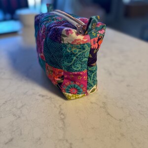 Op de afbeelding: Een rechthoekige, gewatteerde make-up tas met een patchwork ontwerp. De tas heeft verschillende bloemen- en geometrische patronen in tinten van teal, roze, paars en groen. Een zilveren rits loopt over de bovenkant en er zitten kleine stoffen lussen aan elk uiteinde.